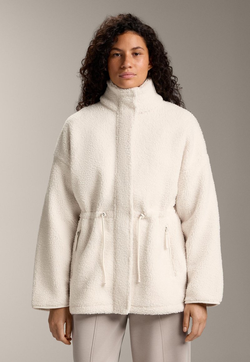 Krém színű polár dzseki magas gallérral, oldalán cipzárokkal és állítható húzózsinórral a deréknál. Dobozos szabású és puha tapintású.