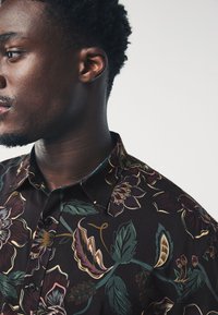 Chemise à manches courtes en tissu foncé avec un motif floral coloré comprenant des verts, rouges et jaunes, col à boutons et coupe décontractée.