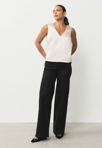 Bez rękawów, kremowy satynowy top z dekoltem w kształcie litery V, zestawiony z luźnymi czarnymi spodniami. Obuwie to czarne buty z ozdobnymi akcentami.