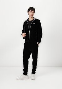 Tuta in corduroy nera con dettagli bianchi, giacca con zip, pantaloni con vita elastica e sneakers bianche. Modello che tiene una borsa nera.