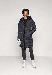 Casaco comprido acolchoado preto com capuz, apresentando fecho de correr frontal, bolsos laterais e textura suave. Usado sobre uma camisola preta e calças.