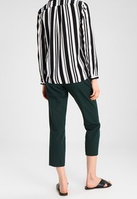 Chemise à manches longues rayée noir et blanc avec col, associée à un pantalon court vert foncé et des sandales noires, vue de dos.