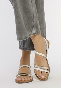 Sandalias blancas con tiras, detalles metálicos, suelas planas y una plantilla texturizada marrón, combinadas con pantalones de mezclilla gris.