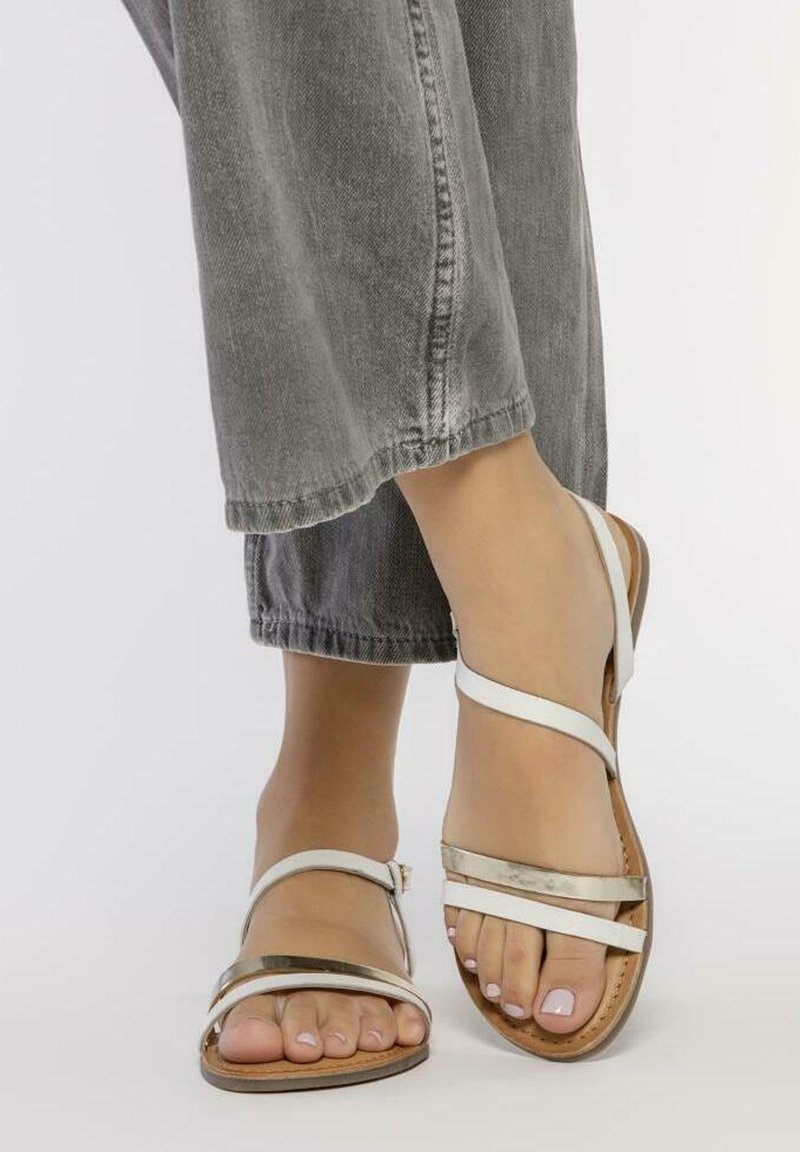 Sandalias blancas con tiras, detalles metálicos, suelas planas y una plantilla texturizada marrón, combinadas con pantalones de mezclilla gris.