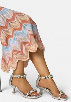 Piernas con sandalias plateadas de tacón bajo con tiras adornadas con piedras preciosas en el tobillo y los dedos, combinadas con una falda multicolor de diseño ondulado.