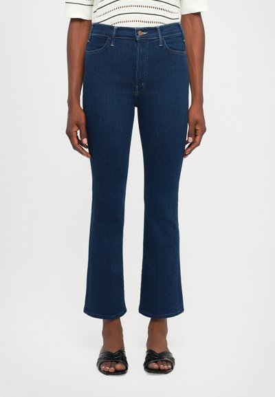 Kvinde iført mørkeblå cropped flare jeans og sorte åbne sandaler, stående mod en enkelt lys baggrund.