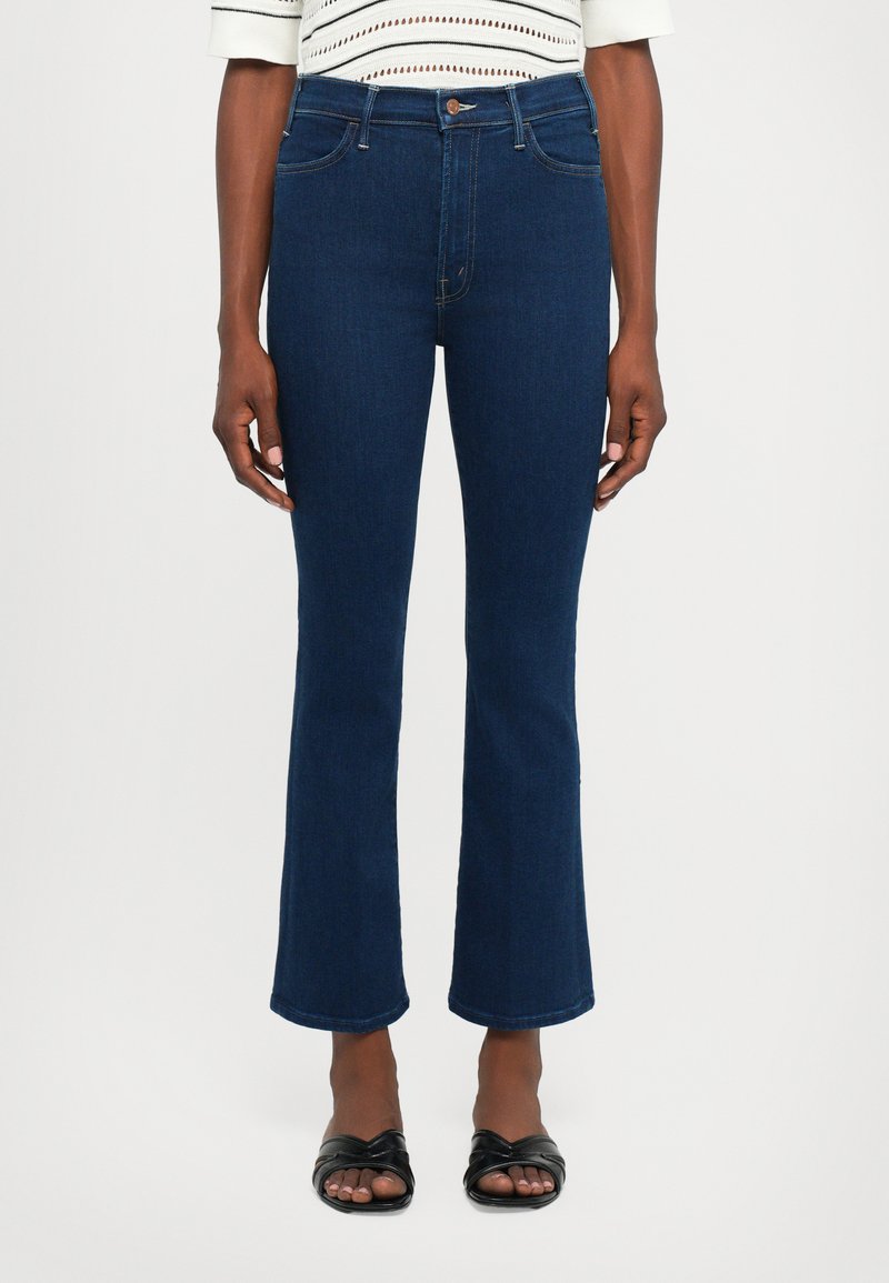Vrouw in donkerblauwe, kortere flare jeans en zwarte open teen sandalen, staande tegen een effen lichte achtergrond.