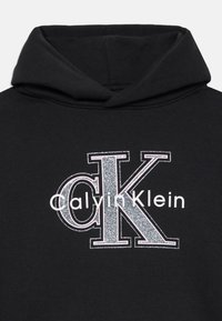 Črna kapuca iz mešanice bombaža, ki vsebuje velik kovinski srebrni logotip z napisom "Calvin Klein". Nežna tekstura in prostorna kapuca.