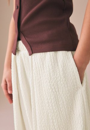 Zbliżenie osoby noszącej brązową, prążkowaną koszulę wpuszczoną w teksturowane białe spodnie, z rękami spoczywającymi przy bokach i w kieszeniach.