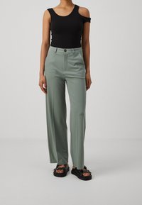 Czarny top na ramiączkach, zielone wysokie spodnie o gładkiej fakturze oraz czarne sandały na platformie z dekoracyjnymi akcentami.