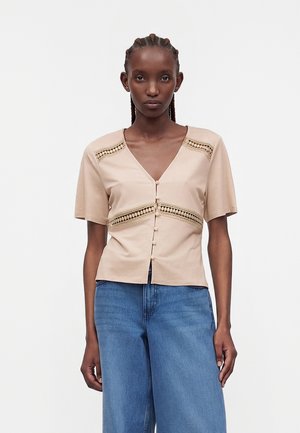 Γυναίκα με πλεξούδες φοράει μια μπεζ κοντομάνικη πουκάμισο με κουμπιά και δαντέλα και μπλε ψηλόμεσο τζιν, στέκεται μπροστά σε λευκό φόντο.