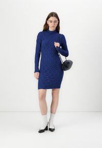 Rochie tricotată albastră cu dungi, cu mâneci lungi și guler înalt, asortată cu o geantă rotundă neagră, șosete albe și tocuri negre din piele lacuită.