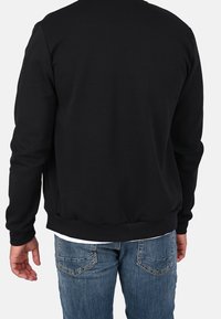 Sudadera negra con cuello redondo, puños y bajo acanalados; parece estar hecha de algodón o una tela similar. Se lleva sobre unos vaqueros azules de mezclilla.