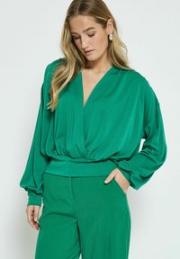 Blusa verde a maniche lunghe con scollo a V profondo e vita elastica, realizzata in un tessuto morbido e fluido. Indossata con pantaloni abbinati.