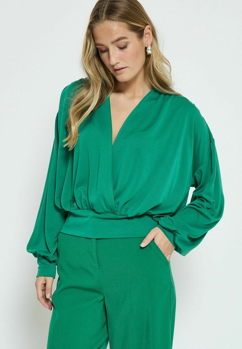 Blusa verde a maniche lunghe con scollo a V profondo e vita elastica, realizzata in un tessuto morbido e fluido. Indossata con pantaloni abbinati.