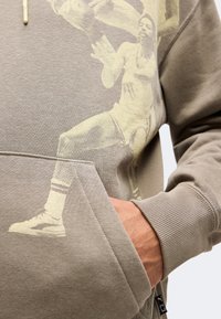 Personne portant un sweat à capuche marron avec une image imprimée d'un joueur de basketball en action à l'avant, la main partiellement dans la poche avant.