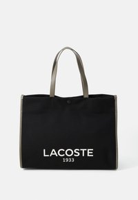 Černá plátěná taška s šedými uchy, na přední straně se bílým logem "LACOSTE 1933" a s patentním uzávěrem na vrchu.