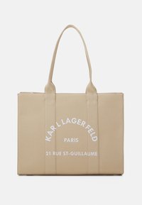 Sac fourre-tout en toile beige avec deux poignées, orné d'un texte blanc indiquant "KARL LAGERFELD PARIS 21 RUE ST-GUILLAUME" dans un design incurvé.
