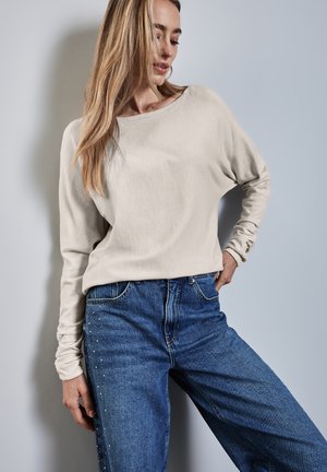 Haut beige clair à manches longues avec un décolleté large, associé à un jean en denim bleu taille haute avec des embellissements sur les côtés.