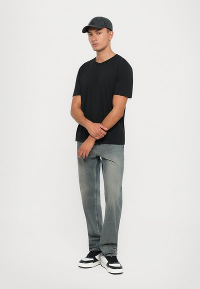 Svart kortermet t-skjorte, lys grå rettbente jeans, svart og hvit sneakers, og en svart caps, stående mot en hvit bakgrunn.