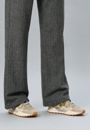 Jambes portant un pantalon en chevron gris et des baskets beige, fauve et vert olive avec des semelles en caoutchouc texturées sur un fond clair.