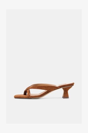 Brūna zamšādas sandalete ar atvērtu purngalu, zemu papēdi un krusteniskām siksniņām, attēlota uz vienkārša gaiša fona.