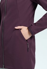 Giacca softshell viola con chiusura a zip, tasche laterali e polsini in velcro; presenta una texture liscia e una vestibilità su misura.