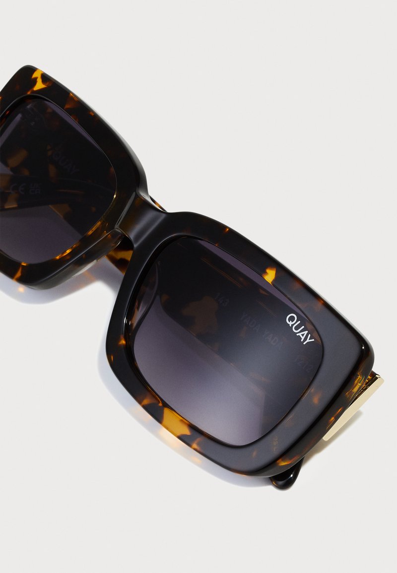 Schildpatt-Sonnenbrille mit einem massiven quadratischen Rahmen, dunkel getönten Gläsern, goldenen Akzenten und dem Markennamen "QUAY" an der Seite.