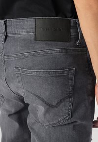 Vaqueros de mezclilla gris con una etiqueta de cuero negro en la cinturilla. Cuenta con dos bolsillos traseros con detalles de costura, textura suave y un corte tapered.