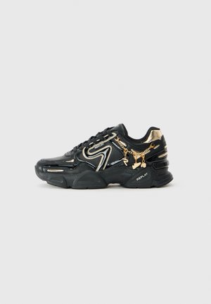 Svart og gull Replay-sneaker med kraftig såle, dekorativ gullkjede og anheng, samt hvite logo-detaljer på siden og hælen.