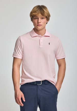 Helles rosa Polohemd aus Baumwolle; mit einem dreiknöpfigen Kragen und kleinem schwarzem Logo auf der Brust. Kombiniert mit dunkelblauen Hosen.