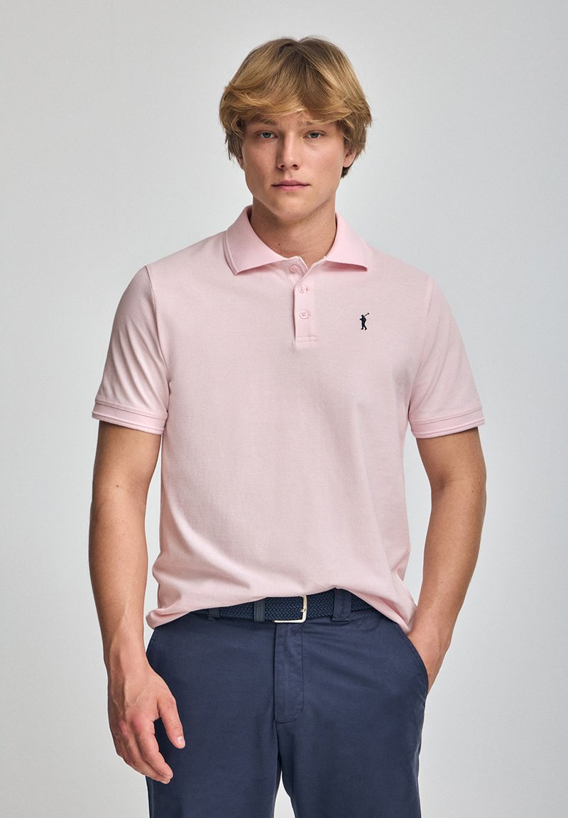 Lyserød polo skjorte lavet af bomuld; den har en krave med tre knapper og et lille sort logo på brystet. Kombination med mørk navy bukser.