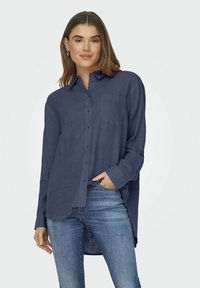 Camicia button-up in lino blu navy con maniche lunghe, una tasca sul petto e orlo curvo, abbinata a jeans azzurri chiari.