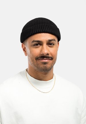 Un bonnet noir à côtes porté avec un t-shirt blanc à col ras du cou. Le t-shirt a une texture lisse, et la personne porte un collier en chaîne en or.