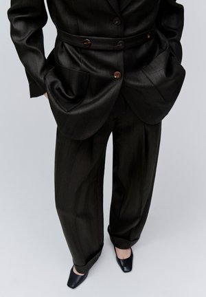 Sötét tűzött mintás öltönyt viselő személy övvel ellátott blézerrel, kezét zsebében tartva, fekete négyzet alakú orrú cipőben, világosszürke háttér előtt áll.