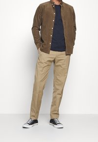 Brązowa koszula z materiału sztruksowego z guzikami, nałożona na granatowy t-shirt, połączona z beżowymi spodniami chinos i czarnymi trampkami z białymi sznurówkami i akcentami.