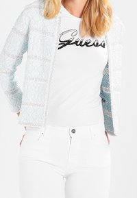 Ljusblå texturerad cardigan med vita detaljer, bärs över en vit t-shirt med svart "Guess"-logotyp, ihop med vita byxor.