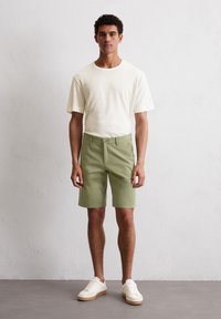 Light beige short-sleeve t-shirt paired with olive green shorts, featuring front pockets. Model wears white sneakers with beige soles.