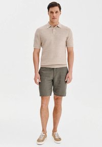 Beżowa dzianinowa koszulka polo z krótkim rękawem, w komplecie z zielonymi szortami. Brązowe trampki dopełniają ten strój, na białym tle.