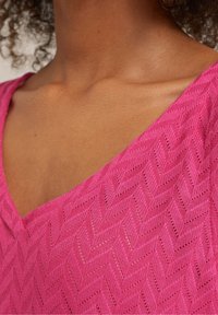 Närbild av ett nacke med mörk hy och lockigt hår som bär en ljusrosa V-ringad topp med ett strukturerat zigzag-mönster.