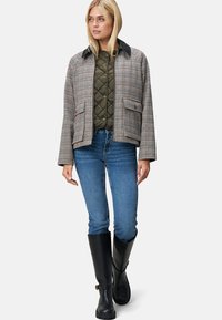 Jasje met houndstoothpatroon in beige, bruin en zwart met een zwarte kraag. Draagt een groene gewatteerde binnenlaag en is gecombineerd met blauwe jeans en zwarte laarzen.