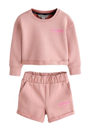 Bambino piccolo con capelli scuri raccolti in codini, che indossa una felpa rosa e pantaloncini, scarpe da ginnastica bianche e calzini bianchi con scritte rosa.
