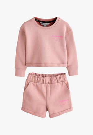 Lille barn med mørkt hår i to fletninger iført en pink sweatshirt og shorts, hvide sneakers og hvide sokker med pink skrift.