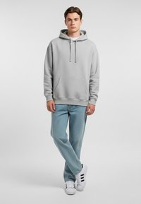 Felpa grigia con tasca frontale e cordini, abbinata a jeans azzurri e sneakers bianche con strisce nere.