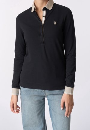 Person trägt ein schwarzes langärmeliges Poloshirt mit beigem Kragen und Manschetten, kombiniert mit hellblauen Jeans, vor einem einfarbigen Hintergrund stehend.