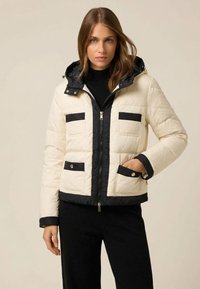 Puffer jacket featuring a cream body with black accents, zip closure, two chest pockets, and a detachable hood. Textured fabric finish.