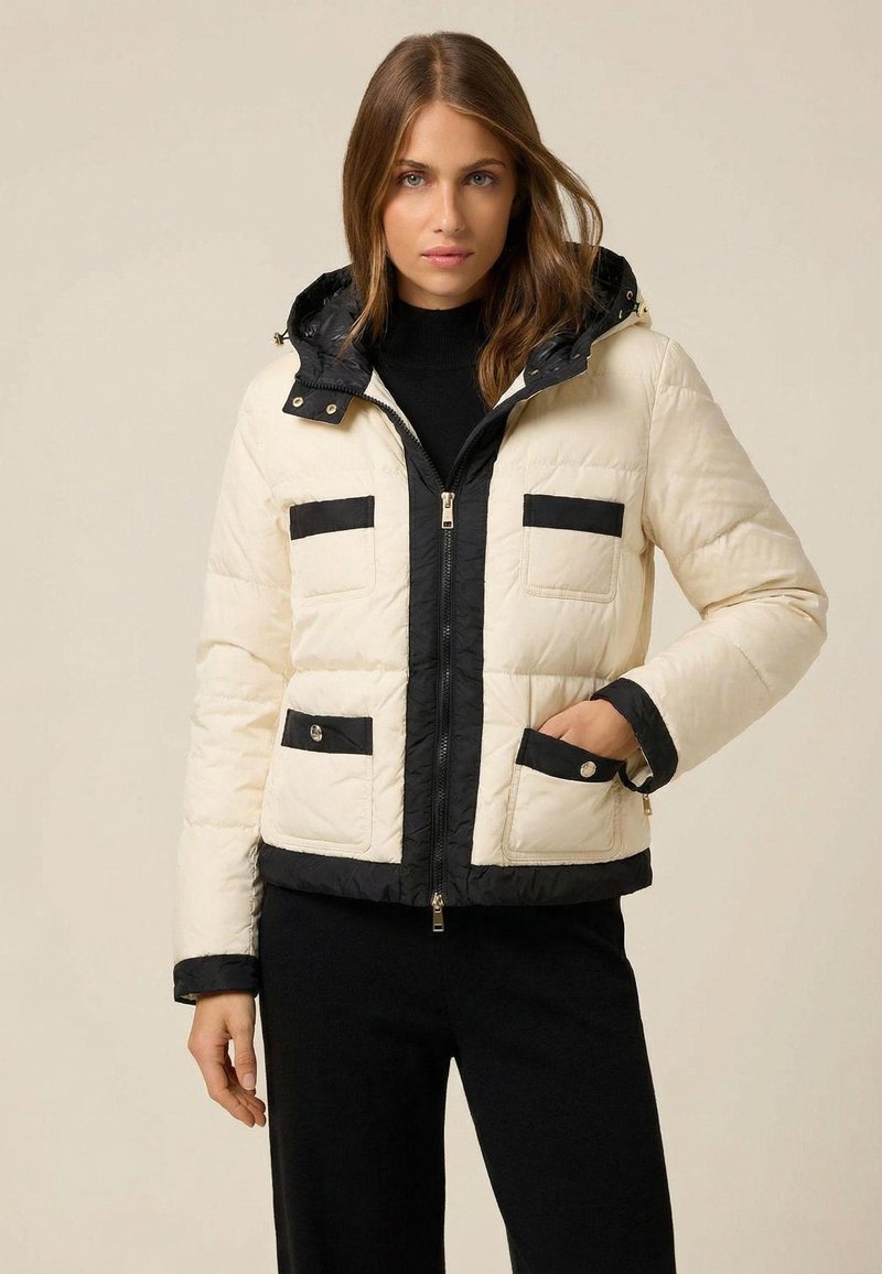 Puffer jacket featuring a cream body with black accents, zip closure, two chest pockets, and a detachable hood. Textured fabric finish.