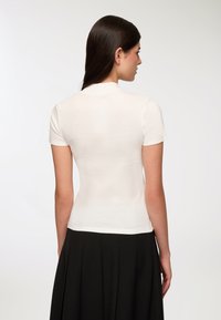 Biały top z krótkim rękawem wykonany z prążkowanego materiału, z wysokim dekoltem i dopasowaną sylwetką. Nosi się go z czarną spódnicą plisowaną.