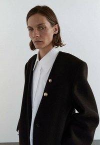 Blazer negro oversized con cuello acanalado, que presenta dos botones dorados en el frente. Debajo, se ve una camisa blanca. Tejido suave y texturizado.