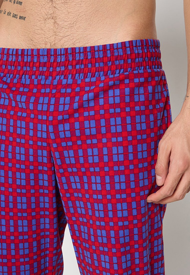 Torso masculino con pantalones elásticos de cintura azul y roja a cuadros, con un tatuaje visible en la parte superior izquierda y la mano derecha descansando cerca del muslo.